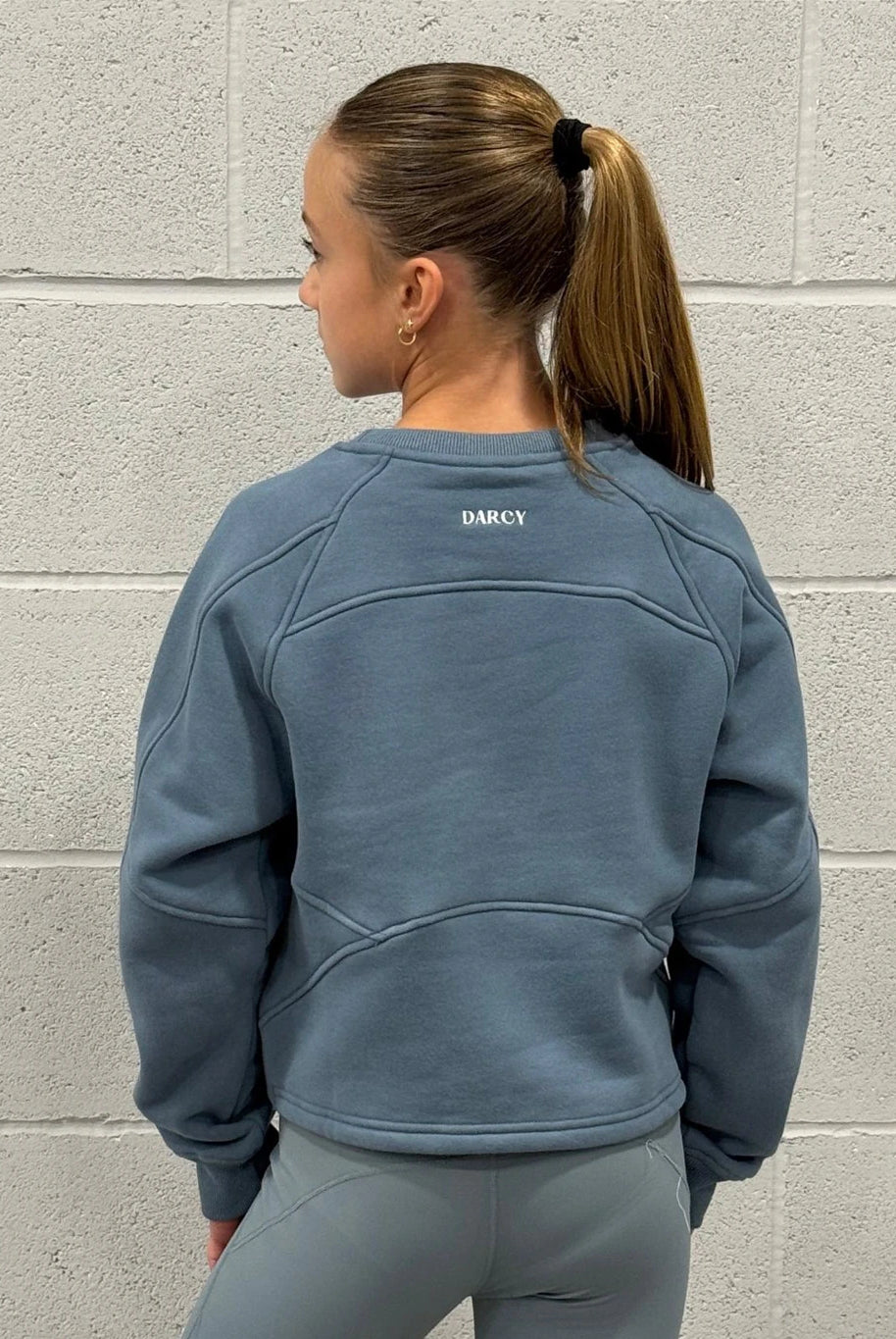 Person wearing a blue sweatshirt with 'Darcy' branding against a gray brick wall.