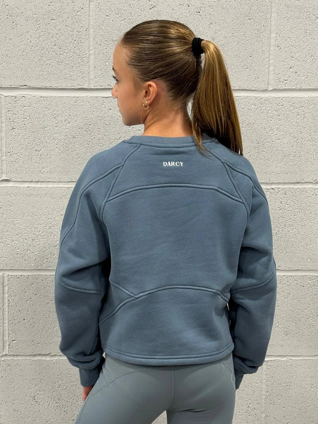 Person wearing a blue sweatshirt with 'Darcy' branding against a gray brick wall.