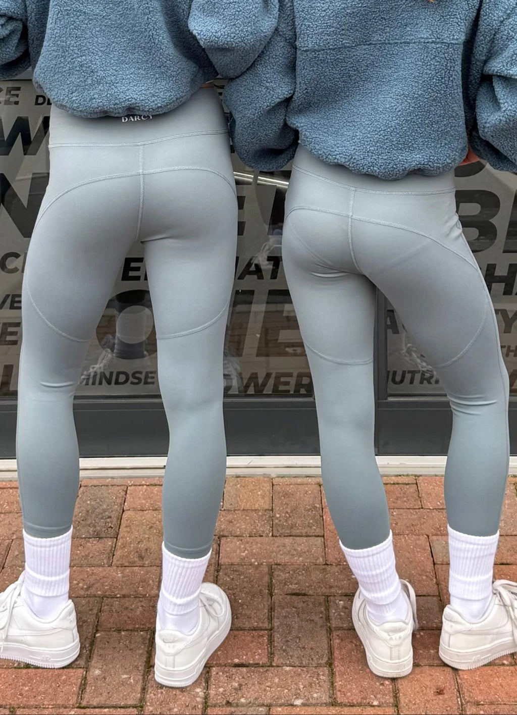 Two people wearing gray leggings and white sneakers on a brick pavement.